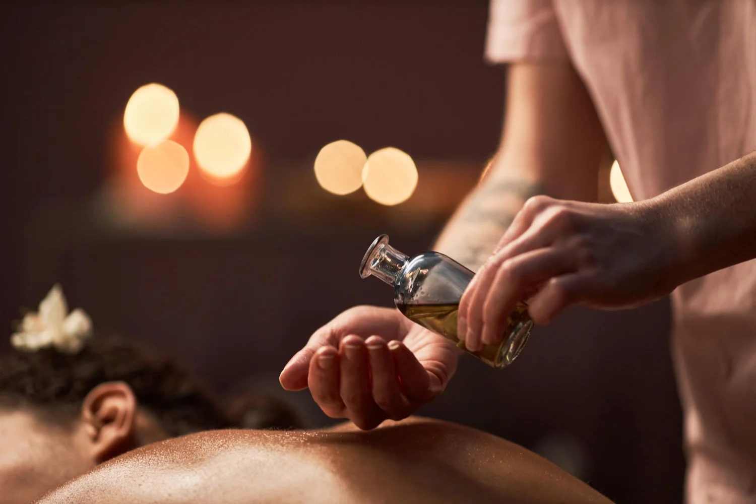Therapist pouring herbal oil onto their palm before applying it to a person's back during a relaxing Ayurvedic massage therapy session, with warm candlelight in the background